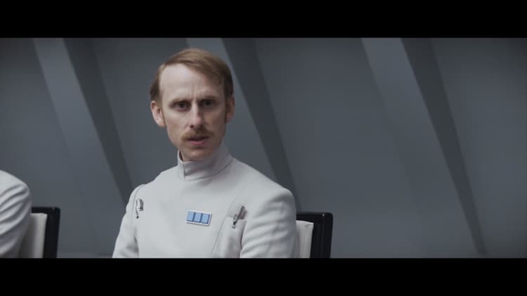An Imperial Security Bureau officer sitting at a conference table looking toward the camera with a confused expression.