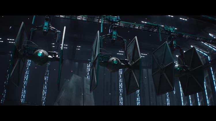 Three Imperial TIE Fighters are lined up side-by-side in a hanger. Pilots are running to enter them and take off.
