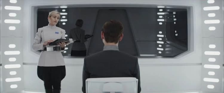 Syril sits in a chair, facing a one-way mirror whilst Meero, standing, holds a data pad while talking to him.