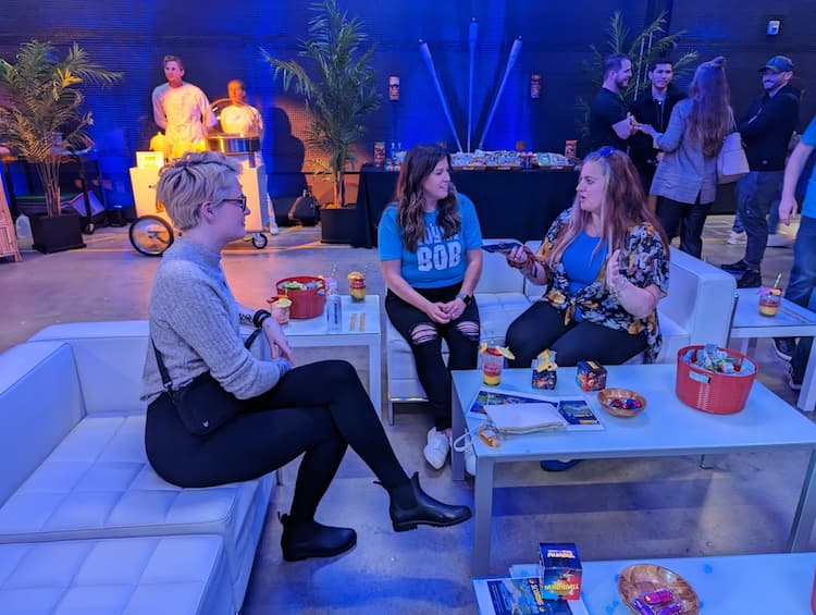 Erika and Michelle sit with Sydney on white couches surrounded by snacks and people.