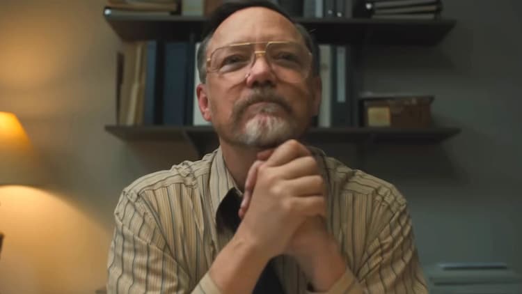 Steve Raglan is sitting at his desk with his hands clasped. Various Books are on shelfs behind him. He's wearing a yellow shirt, glasses, and a purple tie.