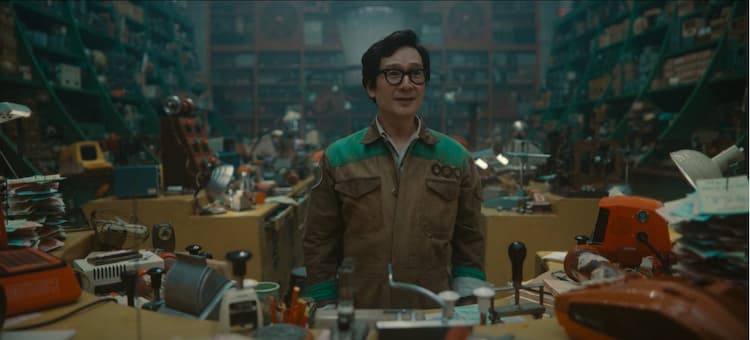 O. B. is standing behind his desk. He is wearing a brown and green TVA uniform. He is surrounded by a variety objects in various states of disrepair.