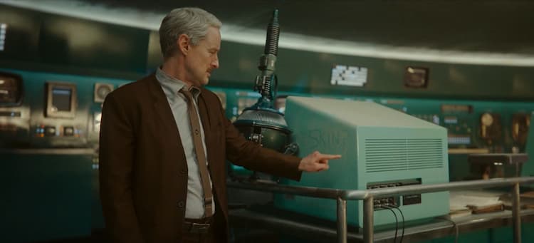 Mobius is standing next to a dusty computer. A device is leaning against the computer and Mobius is writing the word skin with the dust.