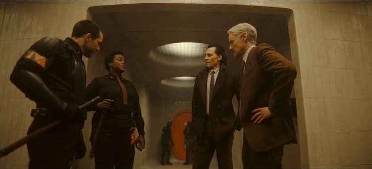 Hunter B-15, Loki, Mobius, and another Hunter are standing outside a holding cell in the TVA. An orange door is down the hall and some light is coming in from above.