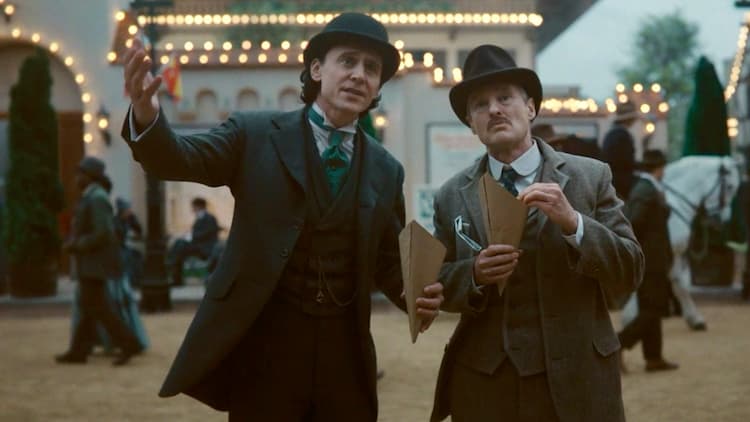 Loki and Mobius are eating snacks. A building covered with lights is behind them. People are walking in both directions behind them as well.