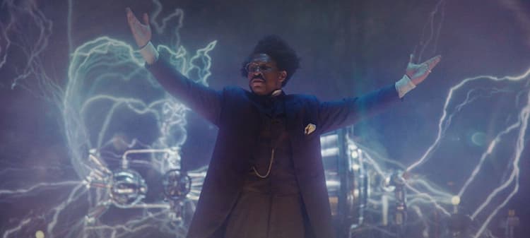 Victor Timely is on a stage giving a presentation. His invention is behind him and the heads of some people can be seen at the bottom of the image.