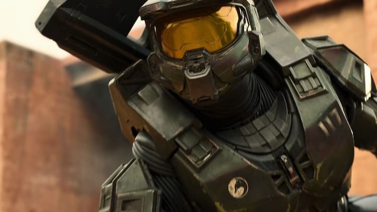 Master Chief is crouched and looking ahead. He has an assault rifle on his bike. The city wall is behind him and the area is covered in red sand.
