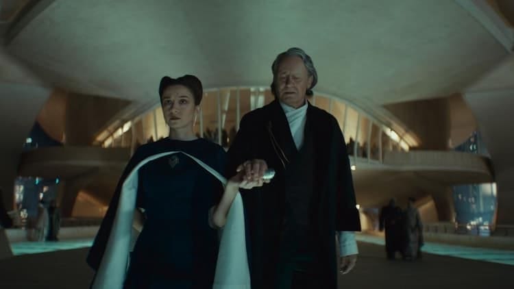 Kleya and Luthen are walking away from an extravagant party. Kleya is handing Luthen an object, and Luthen is grabbing it. Other people at the party are outside as well. It's night. Kleya has a blank look on her face. Luthen has a confused look on his face.