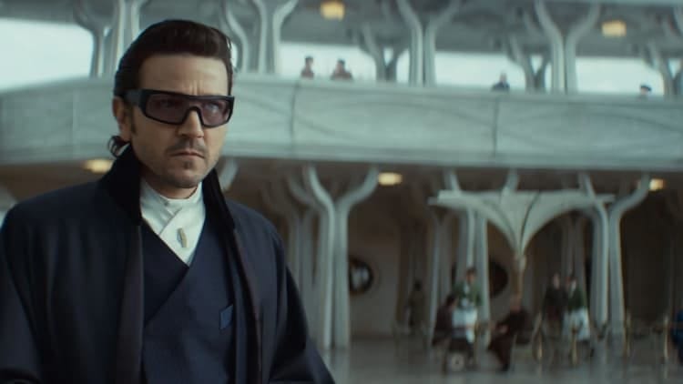 Cassian is in disguise on Ghorman. He is wearing a fashionable jacket with a fancy pair of sunglasses. He is walking through the plaza in the middle of the day. He has a serious look on his face and is looking around the area.