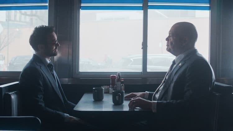 Matt Murdock and Wilson Fisk are sitting in a diner. They are facing each other, and Murdock has his hands underneath the table while Fisk has his hands on top of the table. It's an overcast day outside. They both have serious looks on their faces.