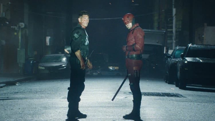 The Punisher and Daredevil are standing in the middle of the street at night. They are looking at the same thing coming towards them. They both look injured. Daredevil is holding his weapon in his right hand.