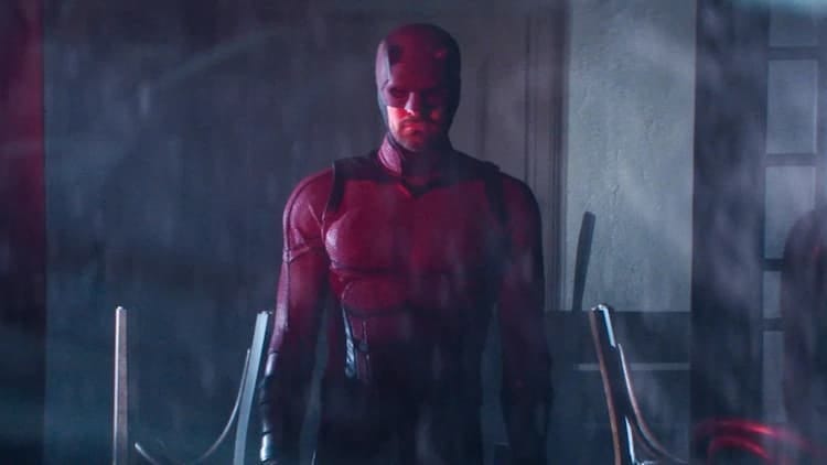 Daredevil is standing in a closed bar. The chairs behind him are upside down on the table. A red light from outside illuminates parts of his body.