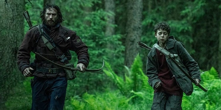 Aaron Taylor-Johnson’s Jamie (bearded, wearing a red raincoat) and Alfie Williams’ Spike (young twelve year-old in a red jumper and faded green coat) run through a verdant forest, holding bows and arrows. Their expressions are serious and winded.
