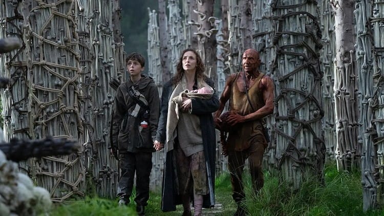 Jodie Comer’s Isla holds Spike’s hand while cradling a baby in her other; Ralph Fiennes’ Kelson, wearing a tank top and covered in something orange leads the other two through tall pillars made of human bones in monument-like structures.
