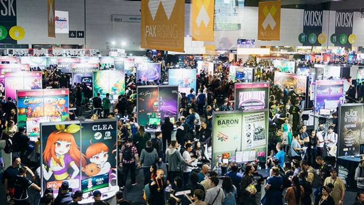 The showfloor at PAX Aus. People are walking around the various booths, looking at and playing games. PAX Aus banners are hanging from the roof.