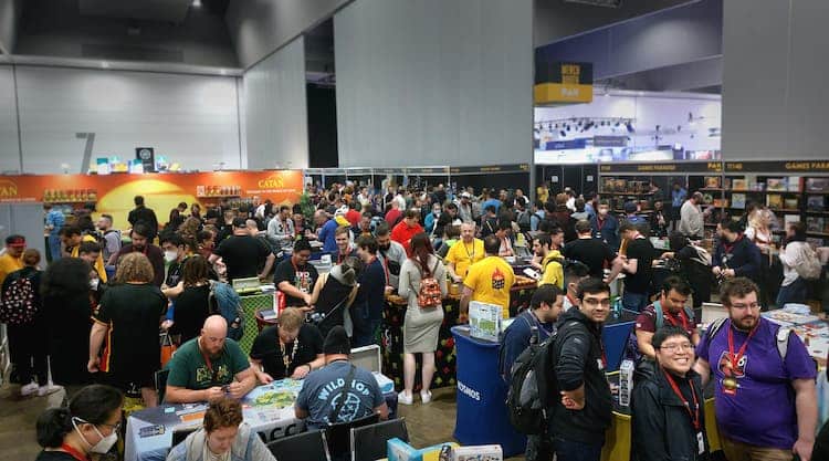 Various people in the tabletop area of PAX Aus. People are playing different games at booths. People are walking through the hall. The expo hall can be seen in the background.