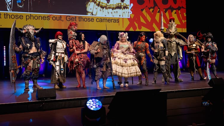 Cosplay participants have lined up on the stage. They are dressed as a variety of different characters. PAX imagery is on the screen behind them.