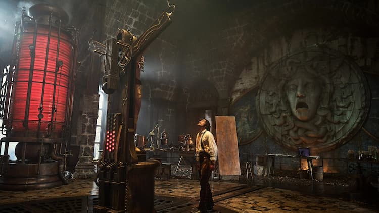 Victor Frankenstein is standing in front of his creation. The Creature is restrained on a cross-shaped table. Victor is looking up at the Creature. Science equipment is scattered around the room. A giant red cylinder is on the left. A face is on the back wall with its mouth open.