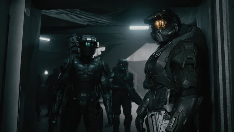 Master Chief and Kai are exploring a facility. Chief is by the door with Kai following him. The area is dimly lit. Another Spartan is in the background.