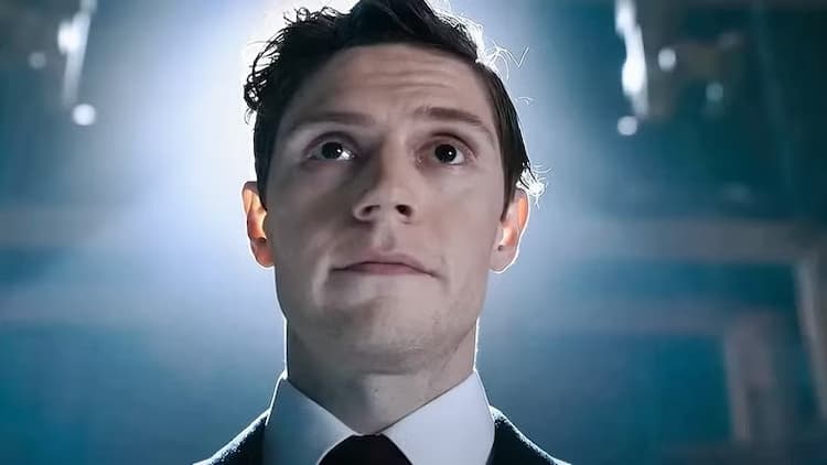 Julian Dillinger is standing in a warehouse with a spotlight on him. He has a confident look on his face. He is wearing a suit and tie.