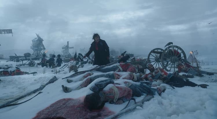 Victor Frankenstein is digging through corpses on a battlefield. The area is covered in snow, corpses, and military equipment. Victor is climbing over multiple dead bodies. His assistants are searching through the other bodies behind him.