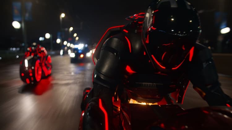 Ares and Athena are riding light cycles down a highway. Both have red accents on their bodies and their light cycles. Normal cars are behind them. Street lights are on. It is night.
