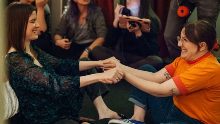 Vic Michalis and Isabella Roland are sitting on the ground, legs crossed, and holding hands. They are smiling and surrounded by crew members.
