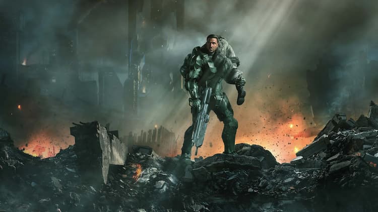 Key art for Season 2 of HALO. Master Chief is standing in the middle of a destroyed city. He has a soldier slung over his shoulder. He is holding a gun in his right hand. The city is burning behind him. He is wearing his armour but does not have his helmet on.