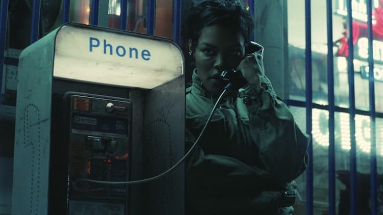 Perfidia is using a payphone at night. She is holding the phone up to her left ear and holding it with her left hand. She has a determined look on her face. The phone box logo is illuminated. Bars are behind her.