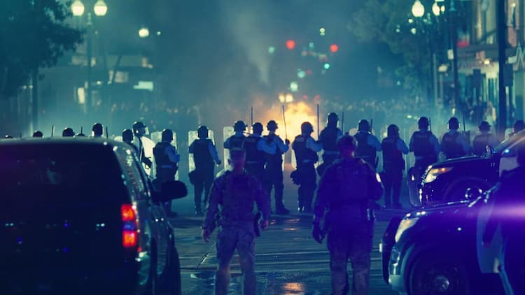 A large group of police and army personnel have set up a wall between them and a protest that they've incited. Lockjaw is at the back of the group, looking towards the protest. A fire is in the distance.