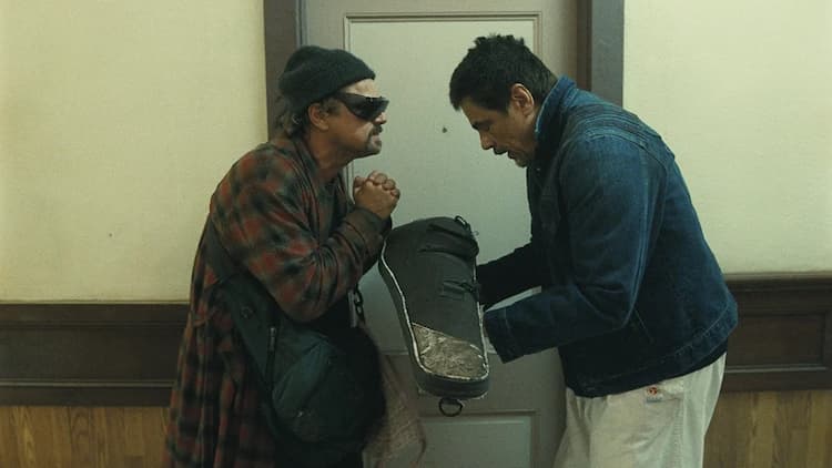 Bob is thanking Carlos in a hallway. Carlos is handing Bob an instrument bag with both hands. Bob is profusely thanking him with his hands clasped. Bob still has sunglasses on.