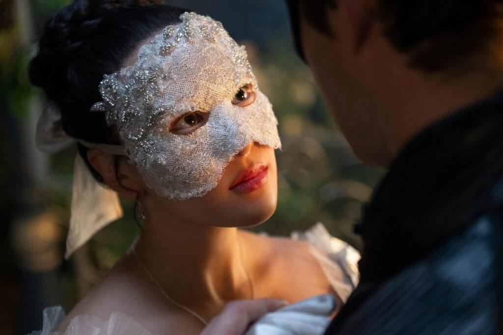 A close-up of a young woman wearing a delicate white lace masquerade mask, looking intently into the face of a man who stands partly out of focus in the foreground. Soft ambient light highlights her features and the ornate texture of the mask.
