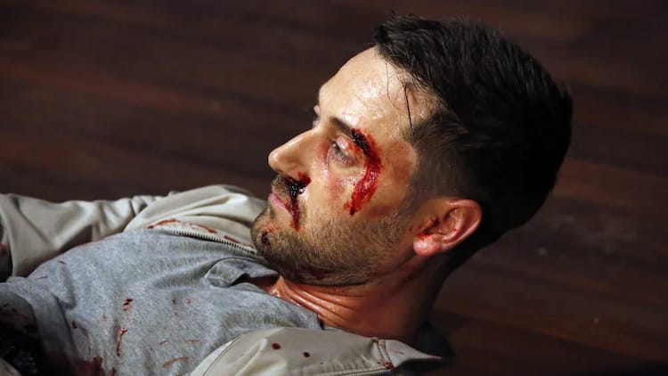 Tom Keen is lying on the floor. He is looking up and left. His face is bloodied.
