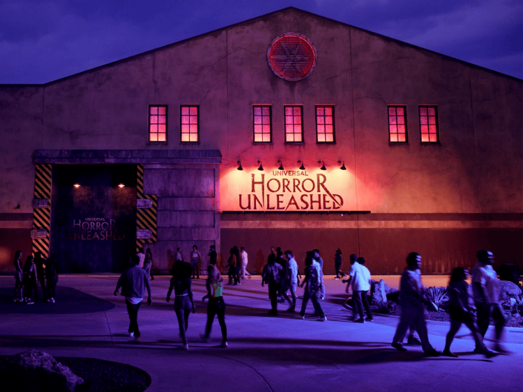 It is nighttime. There is a warehouse that says Universal Horror Unleashed in scary font. There are people walking in front of the warehouse. The colors are tinted red. 