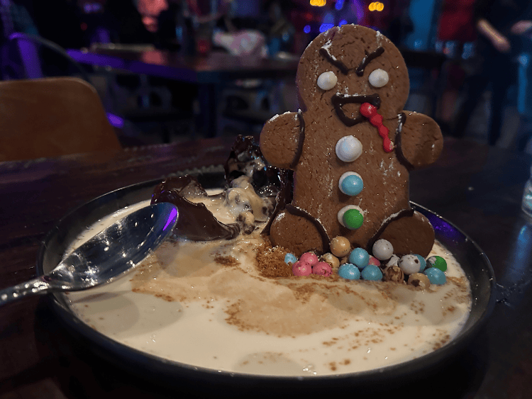 A small black plate holds overflow of pumpkin spice gelato. On the right side of the plate is a large gingerbread man who has angry eyebrows. A semi-melted chocolate dome is behind him. He has several candy buttons at his feet. 