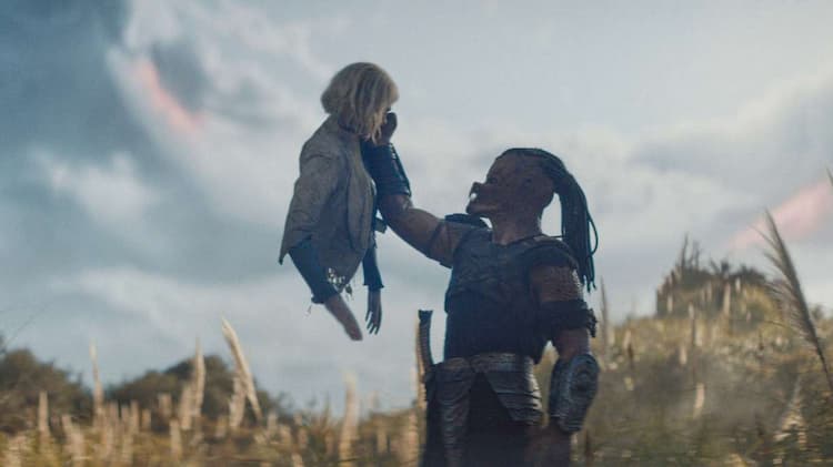 Dek is holding Thia in the air with his right hand. Thia is missing her lower body. Thia is looking down at Dek. They are in the middle of a field.