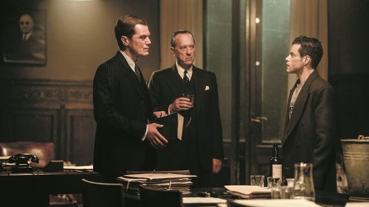 Jackson, Fyfe, and Kelly are in a room together. Jackson and Fyfe are looking at Kelly with confused looks. Kelly is looking at Jackson. It's night outside. Fyfe is holding a glass.