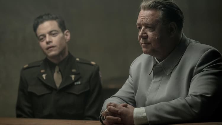 Goring is sitting at a table, his hands clasped. He has a calm look on his face. Kelly is sitting in a chair to Goring's right. He is tilting his head to the right. He is sitting away from the table.