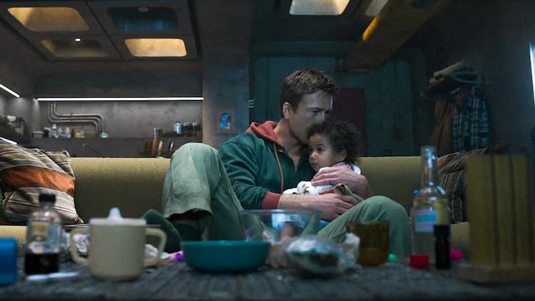 Ben Richards is sitting on a couch with his infant daughter in his arms. Various dishes cover the table in front of the couch. The apartment doesn't have the best lighting. Ben is kissing his daughter on her head.