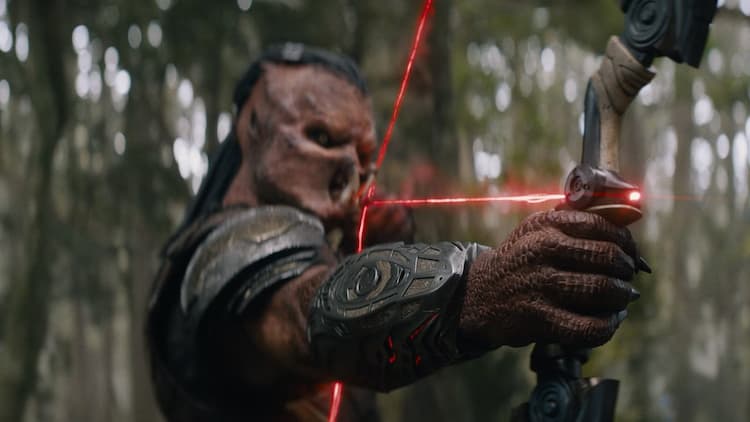 Dek is nocking an energy arrow in his energy bow. The energy is glowing red. Dek has a focused look on his face. He is in the middle of a forest.