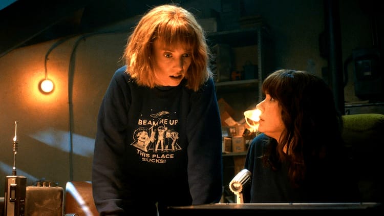 Robin is stood up and hunched over. Joyce is sitting at a desk looking up at Robin.