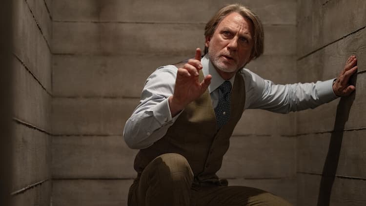 Benoit Blanc is crouched down inside a wooden room. He has his left hand on a wall. He is pointing upwards with his right hand. He has a puzzled look on his face.