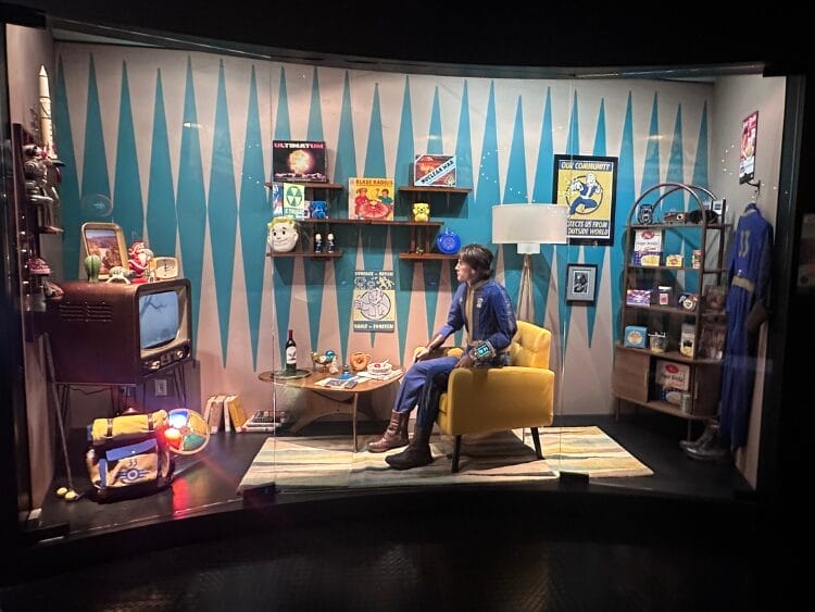 Behind a glass pane is a life-sized set up exhibit of a vault dweller in a wasteland vault. A vault dweller is in their vault dweller suit watching television.
