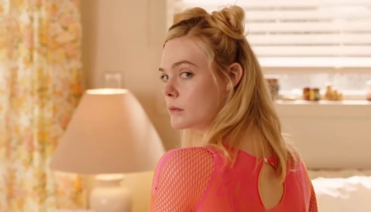 Margot is sitting on a bed. Her body is pointed away from the camera, but she is looking over her left shoulder towards the camera.