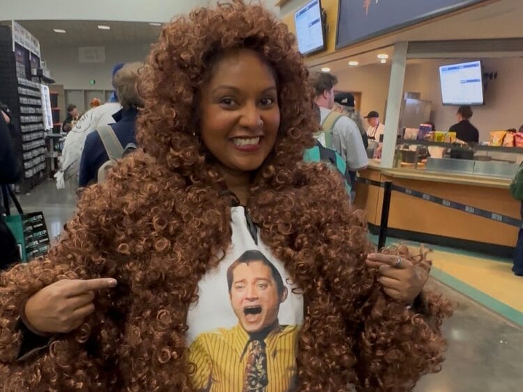 A woman wears a coat of curly brown wigs. She has a shirt with Elijah Wood scream laughing on it. She is smiling.
