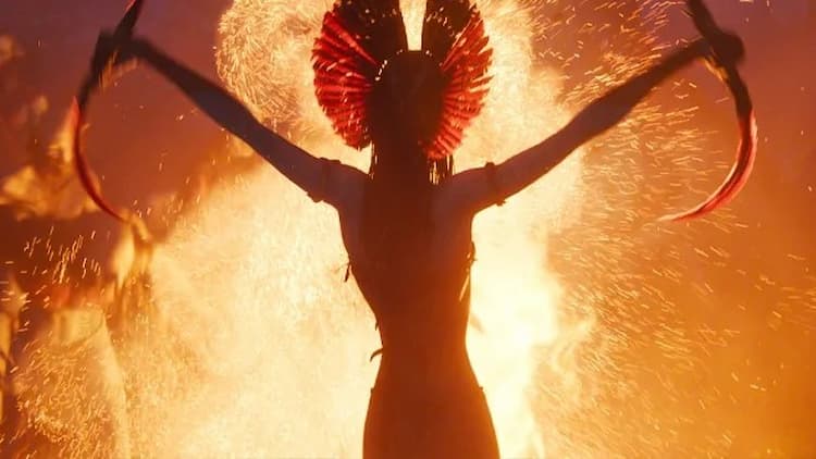 Varang is holding her weapons in front of a fire. She is holding her weapons above her head. She is facing the fire. It's night.