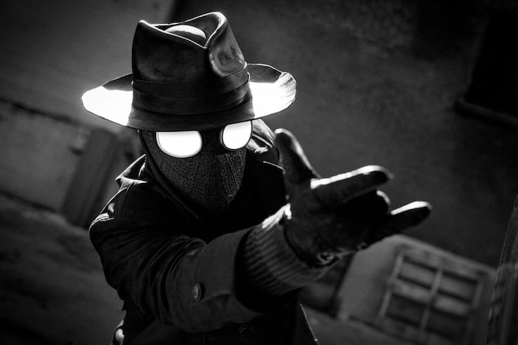 Spider-Man Noir is wearing a gumshoe hat and a long coat. He is pointing his hand like he is shooting webs.