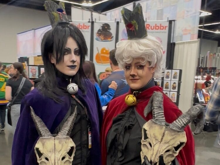 Two cosplayers as the Goat and Lamb from Cult of the lamb. They have large skulls with horns in their arms.