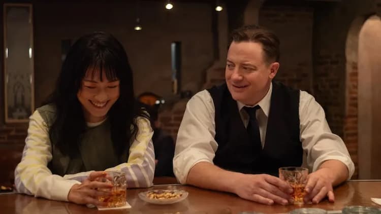 Aiko and Phillip are sitting at a bar. They both have drinks in their hands. Aiko is laughing. Phillip is smiling. The bar is dimly lit.