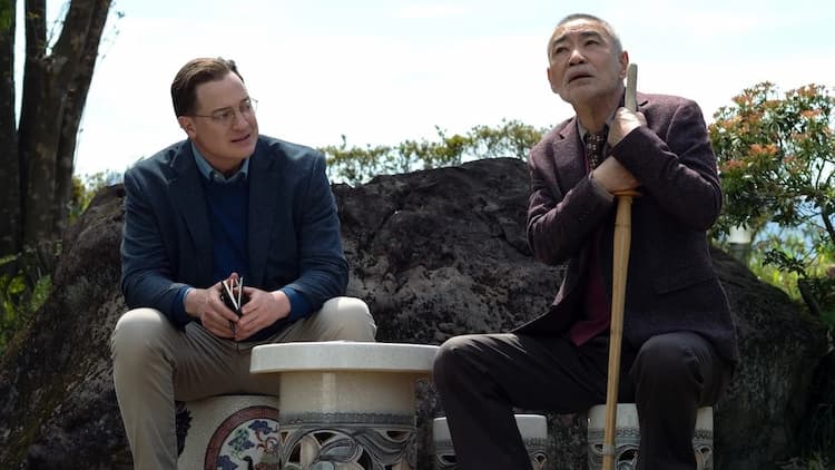 Phillip is listening to Kikuo tell a story. They are sitting at a small table outside. Kikuo is holding a wooden stick. Phillip is holding his notebook. It's the middle of the day.